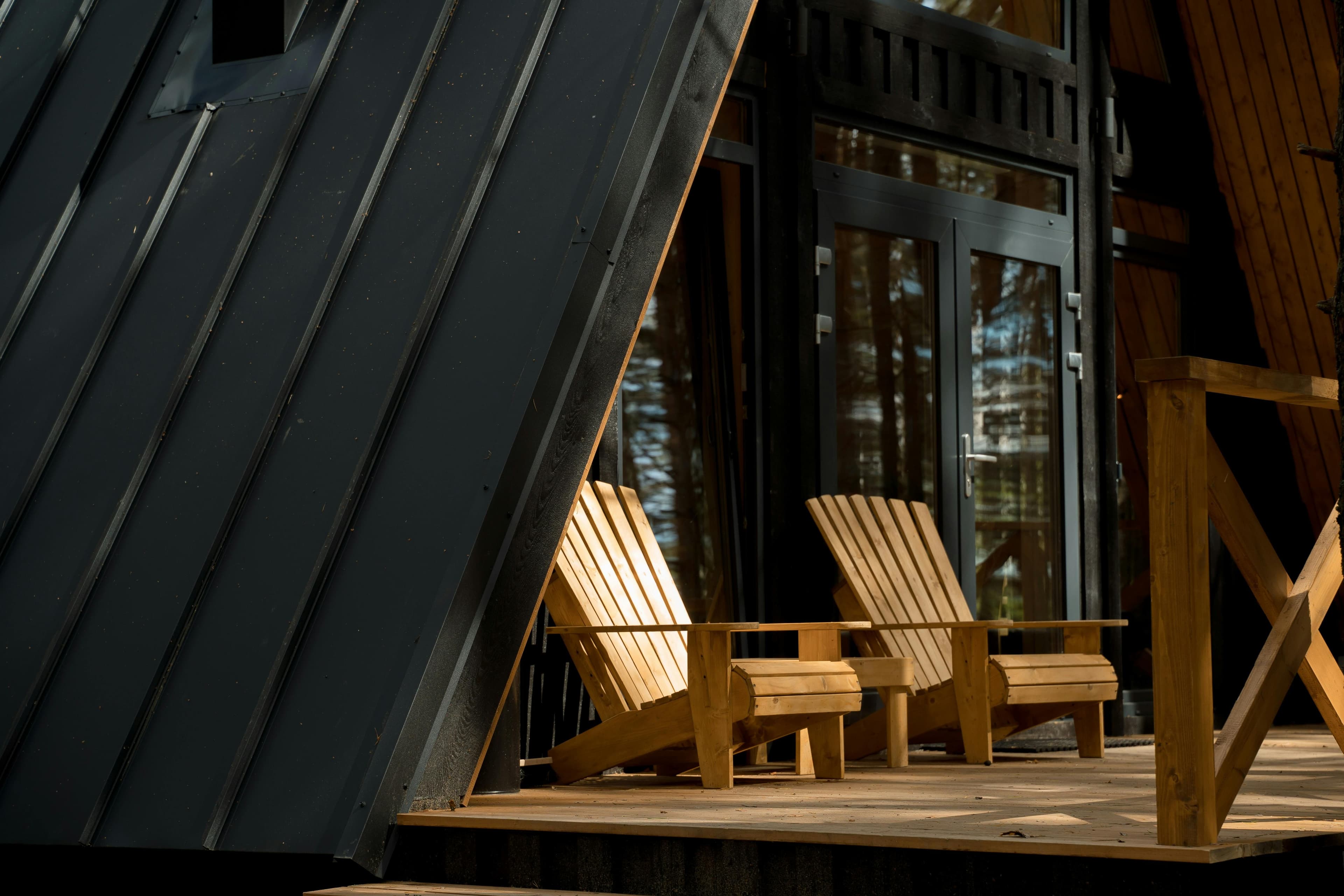 Two wooden Adirondack chairs on a sunlit deck next to a modern dark cabin.