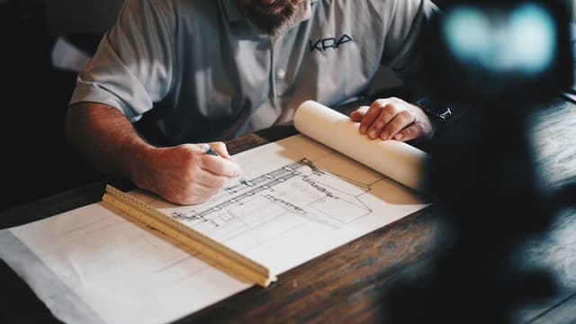 A man wearing a 'KRA' shirt drafts detailed architectural plans with a pen.